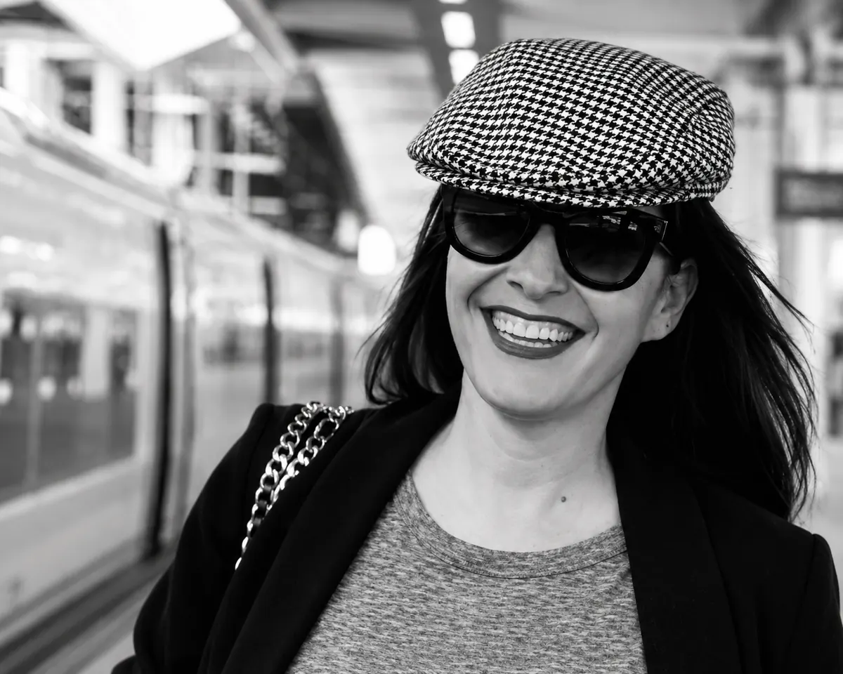 Jessica Bataille sonriente en el andén de una estación, con gorra de pata de gallo y gafas de sol negras